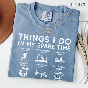 May include: Blue jean Comfort Colors t-shirt with white text: "THINGS I DO IN MY SPARE TIME" and diving-related graphics. Includes a gold necklace with a pendant. The shirt is folded on a white surface.