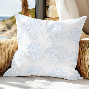 May include: A square white pillow with a repeating pattern of light blue seashell illustrations. The pillow is placed on a wicker chair near a window overlooking the ocean. The pillow is a decorative item.