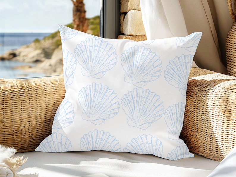 May include: A square white pillow with a repeating pattern of light blue seashell illustrations. The pillow is placed on a wicker chair near a window overlooking the ocean. The pillow is a decorative item.