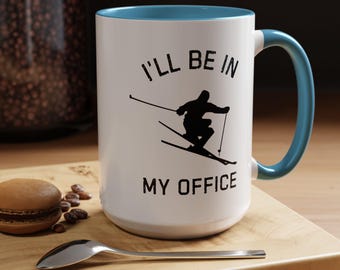 Puede incluir: Taza de cerámica blanca con interior y asa azules. La taza presenta el texto "I'LL BE IN MY OFFICE" con una silueta negra de un esquiador. Un macaron, granos de café y una cuchara están sobre una superficie de madera.