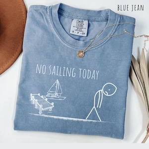 May include: Blue jean-colored t-shirt with the phrase "NO SAILING TODAY" and a graphic of a sailboat and a person. The shirt has a crew neck and short sleeves. A gold necklace with a pendant is draped across the shirt.