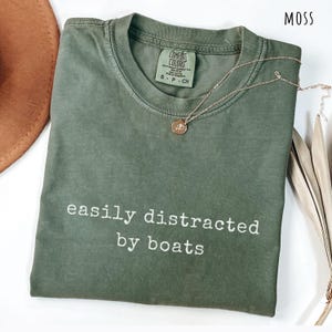 May include: A moss green t-shirt with the text "easily distracted by boats" in white. A gold-colored necklace with a pendant rests on the shirt. The shirt is folded and has a tag that says "Comfort Colors".