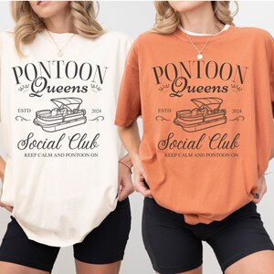 Pu&ograve; includere: Due donne indossano magliette con il testo "Pontoon Queens EstD 2024 Social Club Keep Calm and Pontoon On" e una grafica di una barca ponton. Le magliette sono in avorio e arancio.