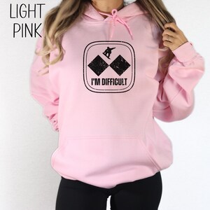 Puede incluir: Sudadera con capucha rosa claro con un gr&aacute;fico negro de un snowboarder en una pista de esqu&iacute; de doble diamante negro. El gr&aacute;fico dice "Soy dif&iacute;cil".