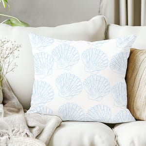 Puede incluir: Cojín cuadrado con un patrón repetido de conchas marinas azul claro sobre un fondo blanco. El cojín está colocado sobre un sofá blanco, con una manta de color neutro y un jarrón de flores en el fondo.