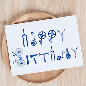 Puede incluir: Una tarjeta de felicitación blanca con las palabras "Happy Birthday" formadas con piezas de bicicleta azules. La tarjeta presenta ilustraciones de componentes de bicicleta como engranajes, cadenas y una bicicleta. La tarjeta está sobre un plato de madera.
