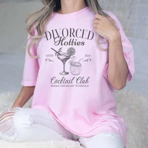 May include: Pink t-shirt with the text "DIVORCED Hotties Cocktail Club Single and Ready to Mingle" and a drawing of two cocktails. The text "ESTD 2024" is also included.
