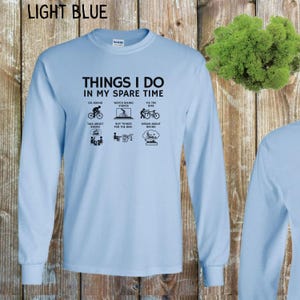May include: Light blue long-sleeved t-shirt with the text "Things I do in my spare time" and a list of activities related to biking. The activities include: go biking, watch biking videos, fix the bike, talk about biking, buy things for the bike, and dream about biking.