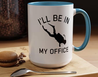Puede incluir: Taza de cerámica blanca con interior y asa azules. La taza presenta el texto "I'LL BE IN MY OFFICE" con la silueta de un buceador. Un macaron, granos de café y una cuchara están sobre una superficie de madera.