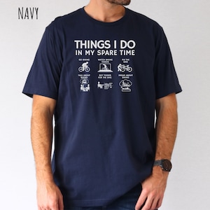 May include: A navy blue t-shirt with a white graphic that says "Things I do in my spare time" and shows icons of a person cycling, watching cycling videos, fixing a bike, talking about cycling, buying things for the bike, and dreaming about cycling.