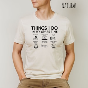 May include: A white t-shirt with black text that reads "Things I do in my spare time."  The text is surrounded by six black icons depicting activities related to cycling.  The word "NATURAL" is printed in black in the top right corner of the image.