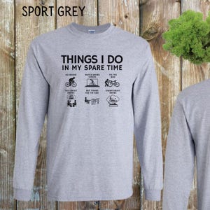 May include: A gray long-sleeve t-shirt with the text "Things I do in my spare time" and illustrations of people biking. The text below the illustrations includes "Go Biking", "Watch Biking Videos", "Fix the Bike", "Talk About Biking", "Buy Things for the Bike", and "Dream About Biking".