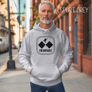 Puede incluir: Una sudadera con capucha gris con un gr&aacute;fico negro de un snowboarder en una pista de esqu&iacute; de doble diamante. El gr&aacute;fico dice "I'm Difficult".