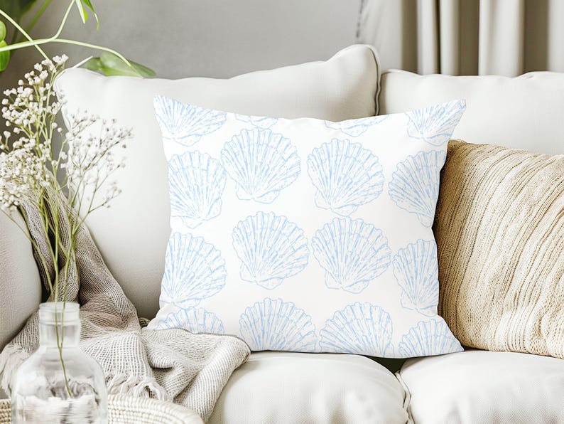 May include: Square throw pillow with a white background and a repeating pattern of light blue seashells. The pillow is placed on a white sofa, with a neutral-colored blanket and a vase of flowers in the background.