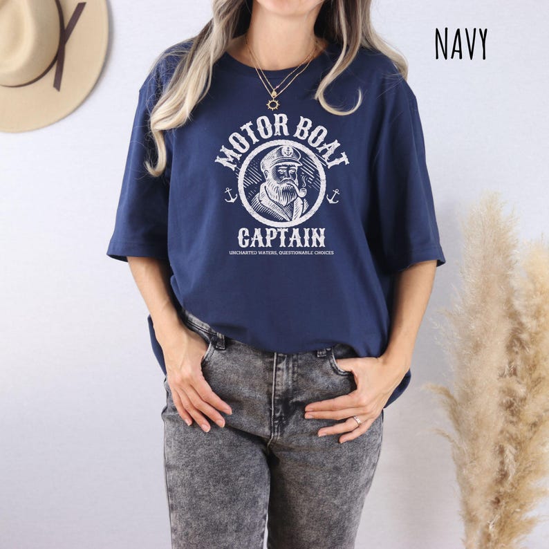 May include: A navy blue t-shirt with a white graphic of a cartoon sailor with the text "Motor Boat Captain" and the words "Uncharted Waters, Questionable Choices".