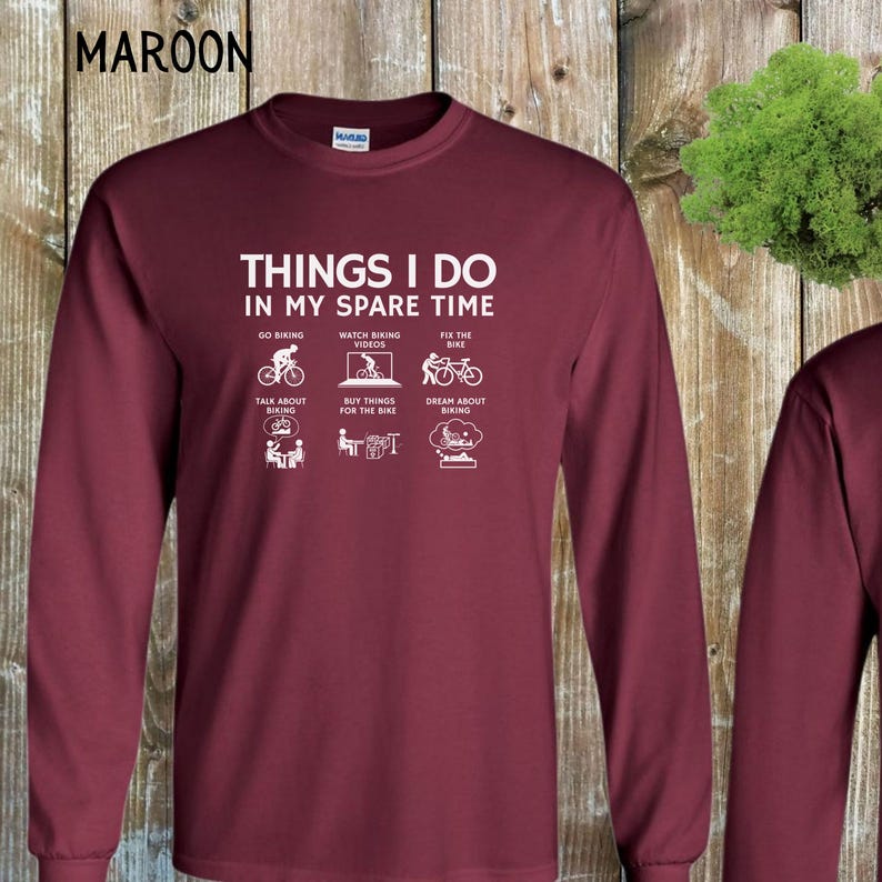 May include: A maroon long-sleeve t-shirt with white text that reads "Things I do in my spare time" and depicts a series of icons related to biking. The icons include going biking, watching biking videos, fixing the bike, talking about biking, buying things for the bike, and dreaming about biking.