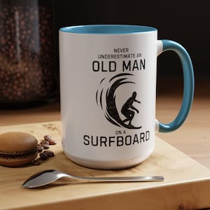 以下が含まれることがあります： 青い内側とハンドルが付いた白いセラミックマグ。マグには「NEVER UNDERESTIMATE AN OLD MAN on a SURFBOARD」の文字と、サーファーの黒いグラフィックが描かれています。木製の表面にはスプーンとマカロンがあります。