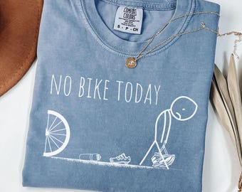 May include: Blue jean-colored t-shirt with the text "NO BIKE TODAY" and a humorous stick figure design. The shirt features a crew neck and a tag that reads "Comfort Colors". A gold necklace with a pendant is draped across the shirt.