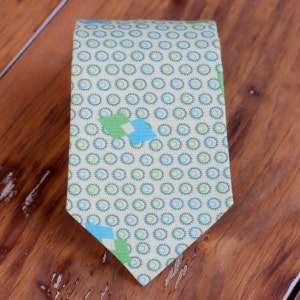 May include: A light green and blue patterned tie with a geometric design. The tie has a light yellow background with blue circles and green and blue diamond shapes.