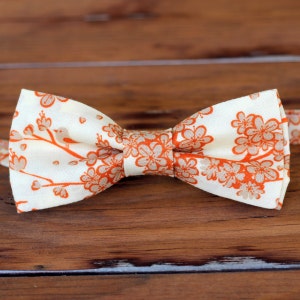 May include: A white bow tie with an orange floral pattern. The bow tie has a small bird design on one side.