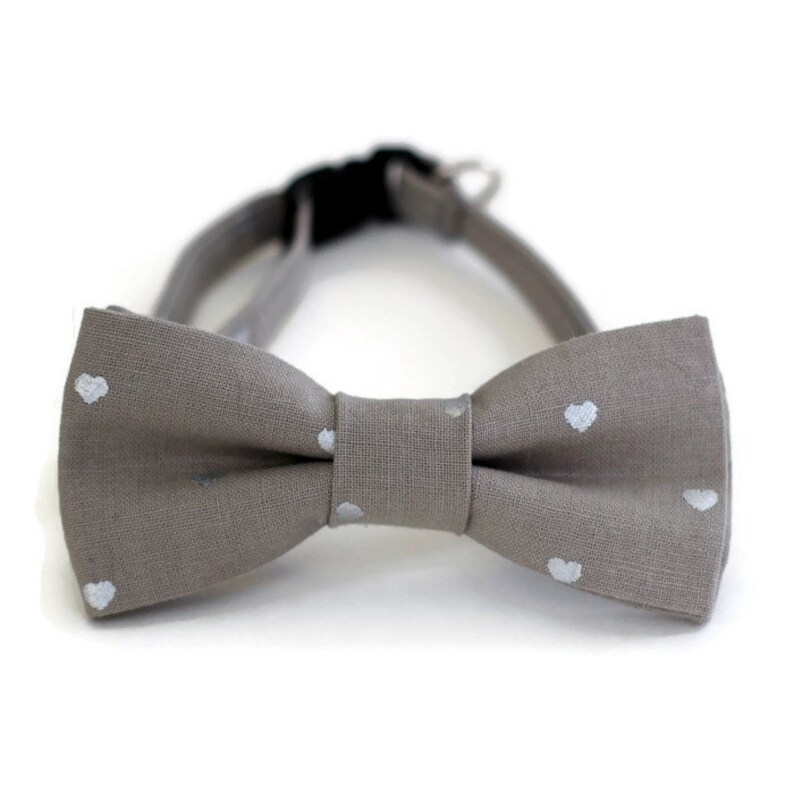 May include: A gray bow tie with white heart-shaped patterns. The bow tie is made of a soft, textured fabric.