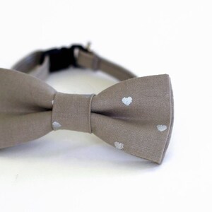 May include: A light brown bow tie with white heart-shaped patterns. The bow tie is made of a soft, textured fabric.