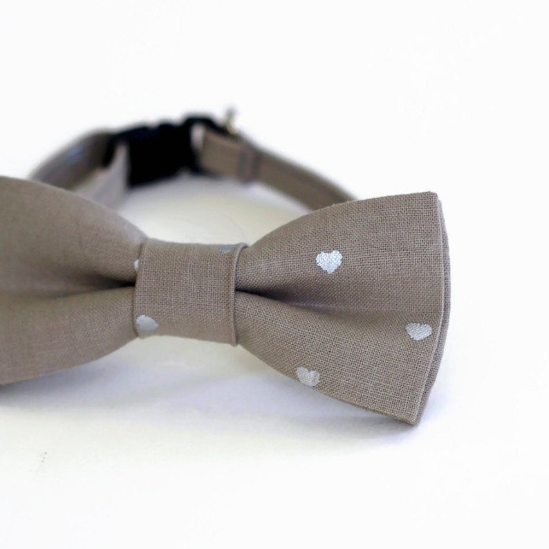 May include: A light brown bow tie with white heart-shaped patterns. The bow tie is made of a soft, textured fabric.