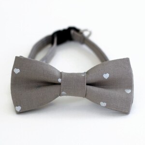 May include: A gray bow tie with white heart-shaped patterns. The bow tie is made of a soft, textured fabric.