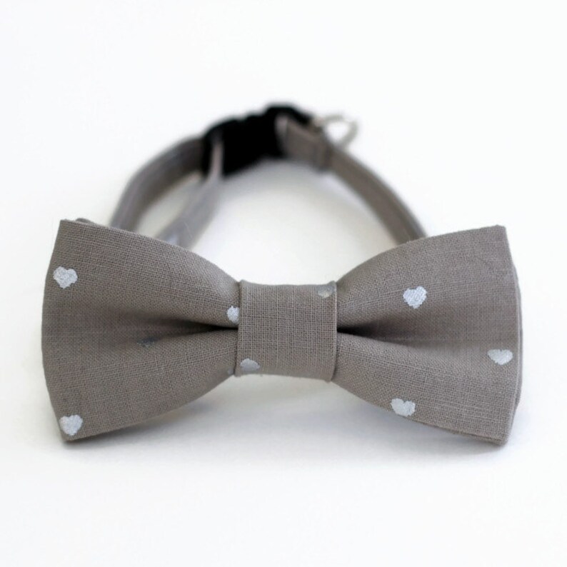 May include: A gray bow tie with white heart-shaped patterns. The bow tie is made of a soft, textured fabric.