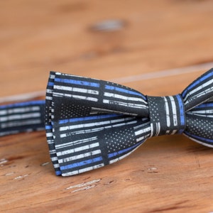 May include: A black bow tie with a thin blue and white stripe pattern. The pattern resembles the American flag with a thin blue line added to represent law enforcement.