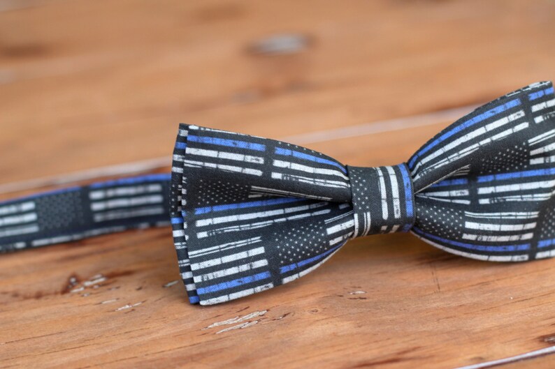 May include: A black bow tie with a thin blue and white stripe pattern. The pattern resembles the American flag with a thin blue line added to represent law enforcement.