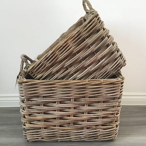 May include: Two woven wicker baskets. The larger basket is rectangular with a square top and two handles. A smaller, oval-shaped basket with a handle sits inside. The baskets are a natural, light brown colour.