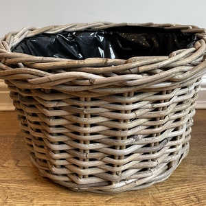 May include: A round wicker planter with a grey wash finish. The planter has a black plastic liner inside.