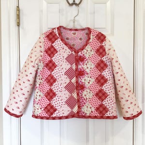 May include: A long-sleeve quilted jacket featuring a patchwork design of red and pink fabrics with floral, gingham, and polka dot patterns. The jacket has a V-neckline, a front button closure with three red buttons, and a light pink floral lining.
