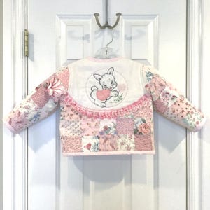 May include: A patchwork jacket in shades of pink, featuring floral and patterned fabrics. The back has an embroidered bunny holding a heart. The jacket has a scalloped lace trim and is hanging on a hanger.