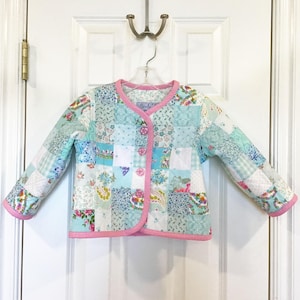 May include: A patchwork jacket with a floral pattern in shades of blue, white, and pink. The jacket has a round collar and is trimmed with pink piping. It features three pink buttons and is hanging on a hanger.
