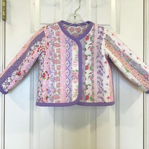 May include: A quilted jacket in shades of pink, lavender, and white, featuring floral and patterned fabric strips. The jacket has a round collar, long sleeves, and is secured with three buttons. The trim is a solid lavender color.