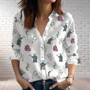 Könnte beinhalten: Weißes Button-Down-Hemd mit einem sich wiederholenden Muster von Cartoonfiguren in verschiedenen Farben. Das langärmlige Hemd hat einen Kragen und ist aus einem leichten Stoff gefertigt. Zu den Figuren gehören eine blauhäutige Figur, eine rosa Figur und eine grüne Figur.