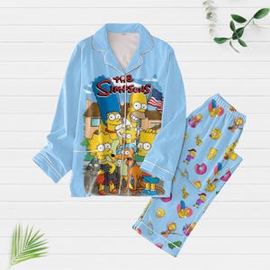 Könnte beinhalten: Hellblaues Pyjama-Set mit einem Simpsons-Motiv. Das Oberteil hat ein Knopfdesign mit weißem Besatz, und die Hose hat ein sich wiederholendes Cartoon-Muster. Das Set beinhaltet ein langärmeliges Hemd und eine passende Hose.