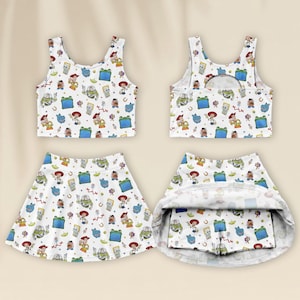 Könnte beinhalten: Ein weißes Sport-Tanktop und ein passender Skort mit einem Allover-Druck von Toy Story-Figuren wie Woody, Buzz Lightyear und Mr. Potato Head. Das Tanktop hat einen Rundhalsausschnitt und der Skort verfügt über eine integrierte kurze Hose.