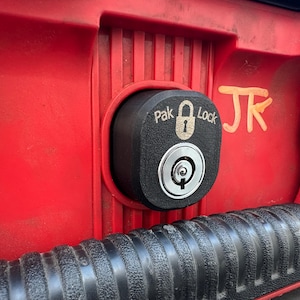 Puede incluir: Cerradura negra Pak Lock instalada en una superficie roja. La cerradura tiene una cerradura y el texto "Pak Lock" impreso. Una manija negra y las letras naranjas "JK" son visibles.