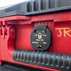 Puede incluir: Primer plano de una caja de herramientas roja y negra con un Pak Lock ML2-DJ Drilling JIG negro. La caja de herramientas tiene un asa negra y una bisagra negra y roja. Las iniciales "JR" están pintadas en naranja.