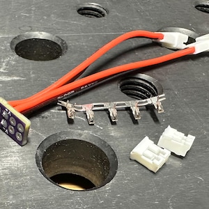 Puede incluir: Un conjunto de componentes electrónicos, que incluyen una placa de circuito con pines, cables rojos y negros, conectores blancos y terminales metálicos. Los componentes están dispuestos sobre una superficie oscura con agujeros circulares.