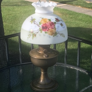 May include: A vintage oil lamp with a brass base and a white glass shade with a floral design. The shade features painted roses in shades of pink, yellow, and red.