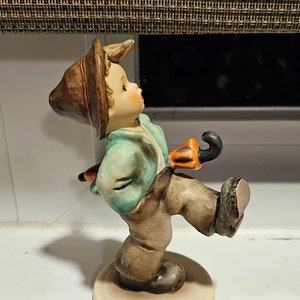May include: A figurine of a boy wearing a brown hat and a blue and green jacket, holding an orange and black umbrella. The figurine is made of ceramic and is standing on a white base.