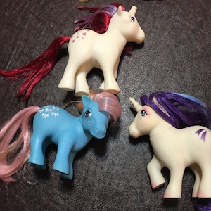 May include: Three My Little Pony toys, one white with a pink and purple mane and tail, one blue with a pink mane and tail, and one white with a purple mane and tail.