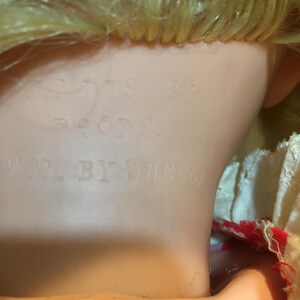Peut inclure: Gros plan sur la nuque et la t&ecirc;te d'une poup&eacute;e vintage. La poup&eacute;e a des cheveux blonds et un teint p&acirc;le. Les mots "DISNEY PROPS" et "MFR. BY UNEEDA" sont estamp&eacute;s sur le cou de la poup&eacute;e, indiquant son fabricant et son origine.