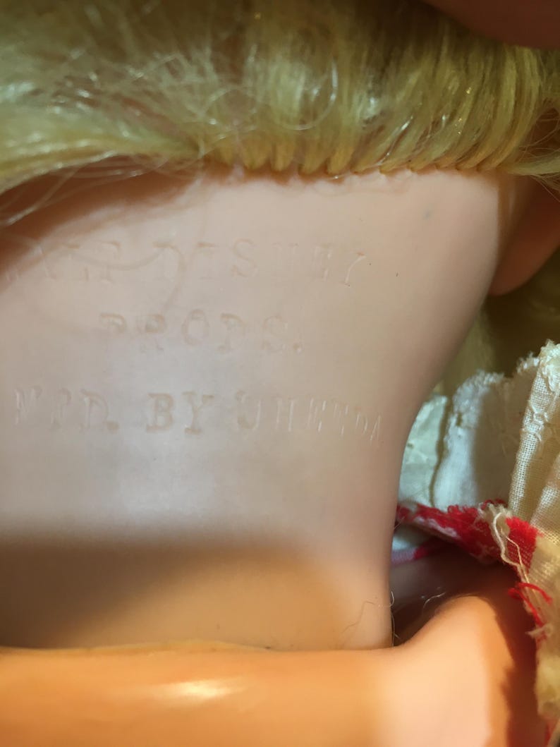 Peut inclure: Gros plan sur la nuque et la t&ecirc;te d'une poup&eacute;e vintage. La poup&eacute;e a des cheveux blonds et un teint p&acirc;le. Les mots "DISNEY PROPS" et "MFR. BY UNEEDA" sont estamp&eacute;s sur le cou de la poup&eacute;e, indiquant son fabricant et son origine.