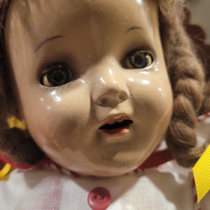 May include: A vintage porcelain doll with brown hair, wearing a white dress with red and white stripes and a yellow ribbon. The doll has a painted face with blue eyes and a red mouth.