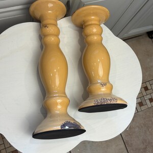 May include: Two glossy, honey-colored ceramic candle holders with a smooth, rounded, turned-wood style silhouette. Each piece features a wide circular base, a slender neck, and a flared top, with visible dark, distressed wear on the edges.